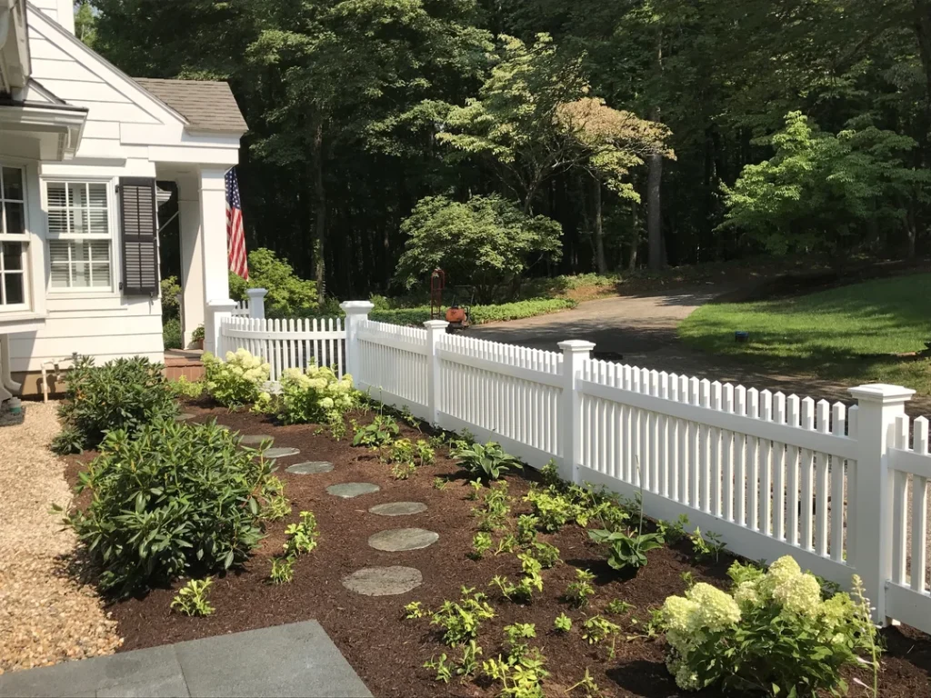 A white house with an American flag, a gravel path, a white picket fence, and a mulched garden bed.